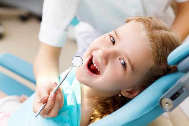 A young girl smiling in the dental chair while a dentist holds a dental mirror
